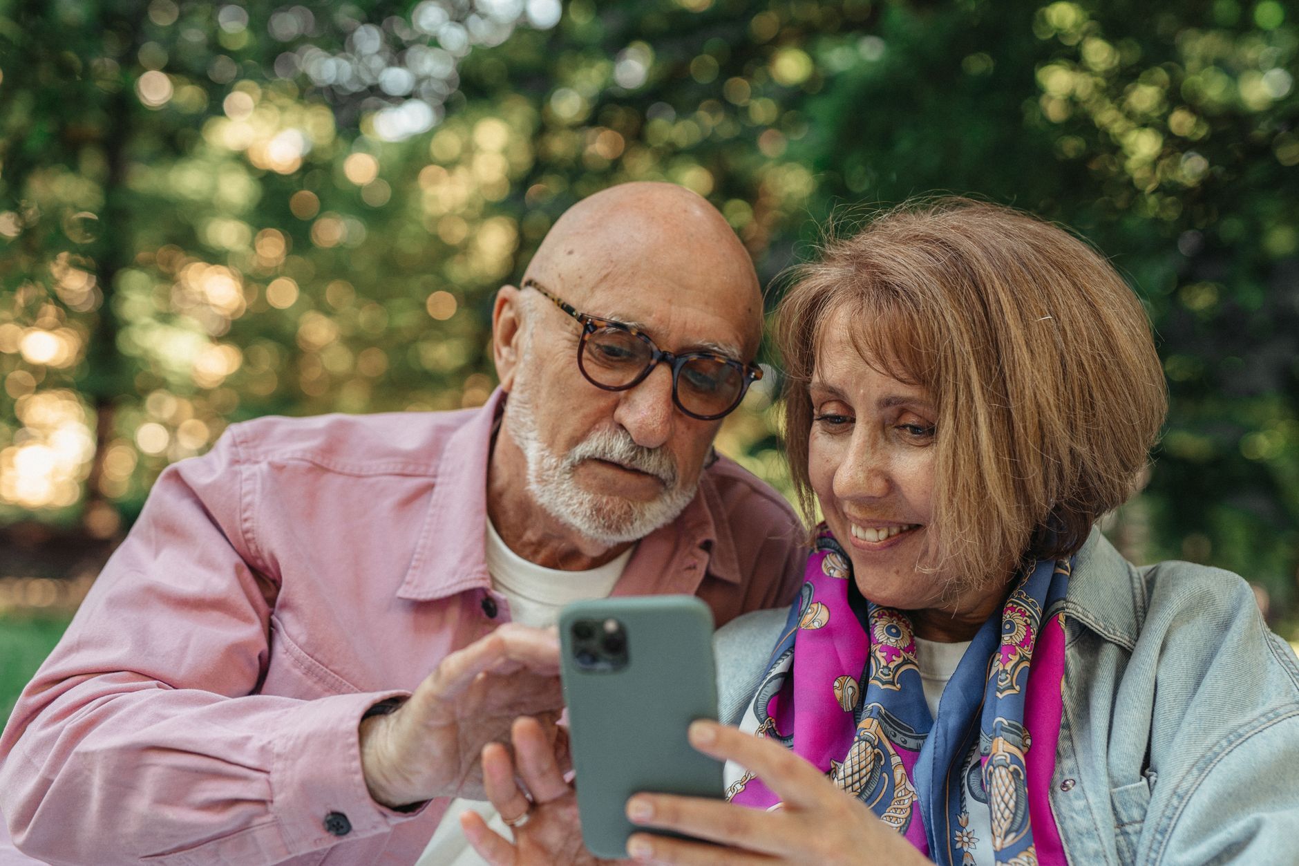 Happy senior couple using smartphone in a park, enjoying sunny outdoors. Photo by SHVETS production on Pexels