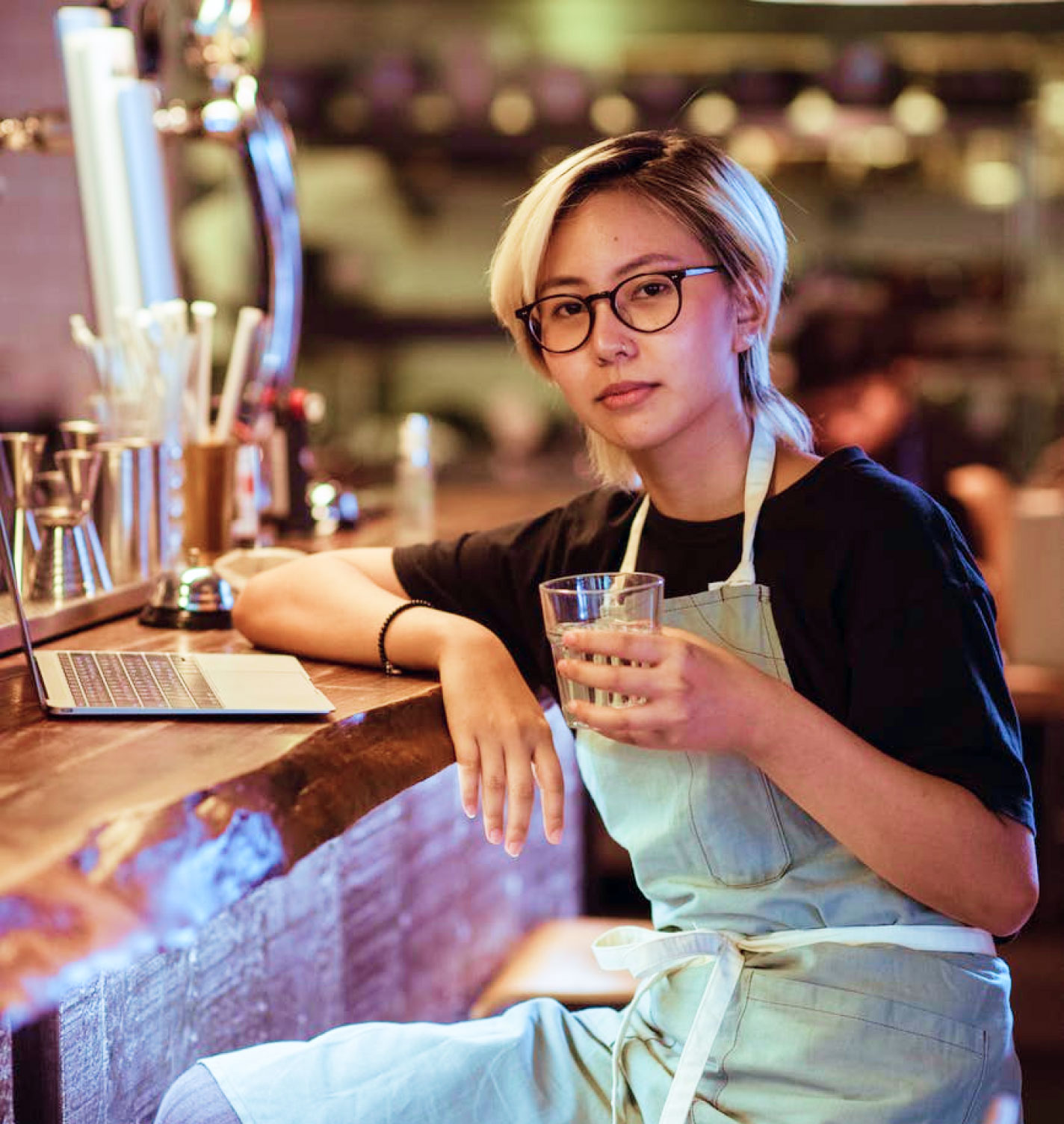 Staged young woman holding glass of water sitting at bar with laptop