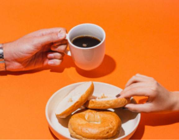 Hands grabbing coffee mug and bagel from centre of image on an orange background