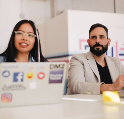 Two coworkers listening to a meeting with laptop covered in stickers on table 