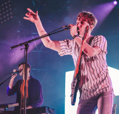 Two men singing on stage with pink lights: one playing keyboard and the other playing guitar